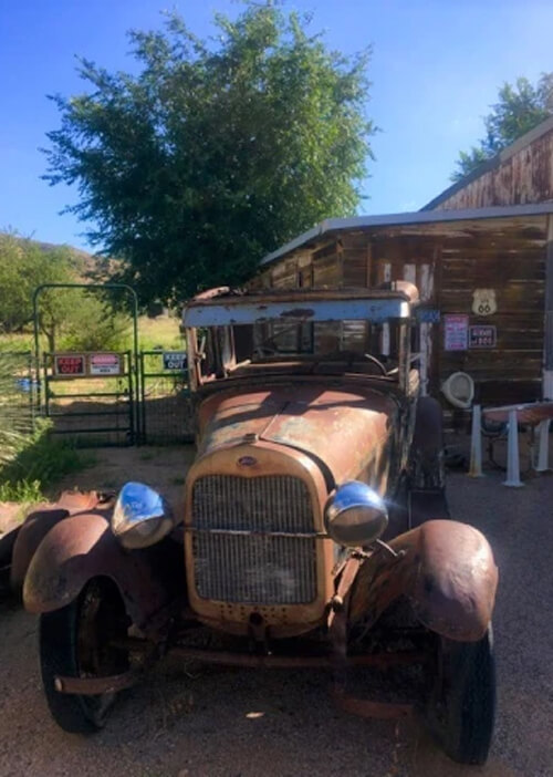 Rusty 1920's Model T Ford filling out bottom of photo, facing strait ahead in front of old wooden store. Green tree and blue sky in background.