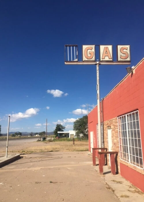 Old sign at right reading "Gas" next to red colored single-story board-up car repair shop on right side, road on left side. Blue sky and a few clouds in background.