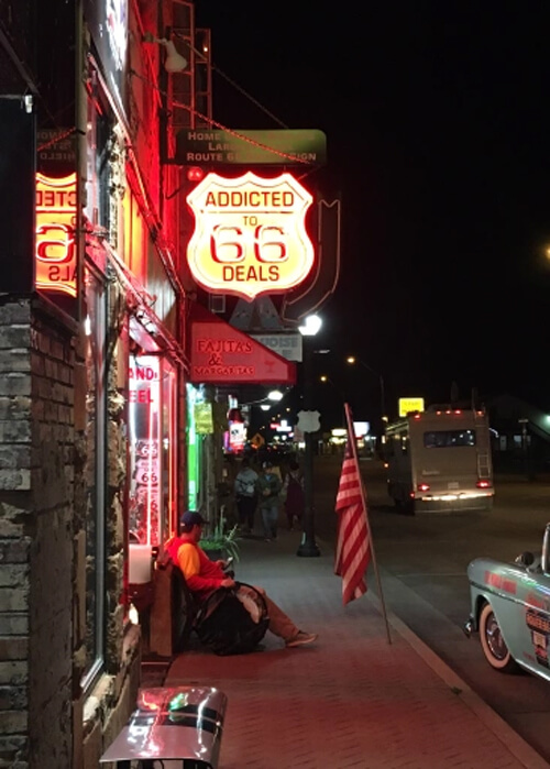 Night photo with lit up "Addicted to Route 66 Deals" sign above store awning at left. A few cars at right on the street, including from of old 1950's car.