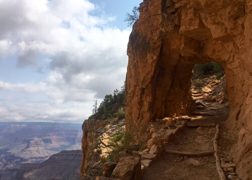 Red rock hiking tunnel at right, with red rock ridges and canyon at bottom left. Blue sky with puffed white clouds in upper left.