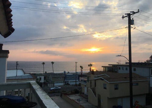 Blotch of sun through white and grey clouds at sunset, with reflection on ocean below, and row of beach homes and palm trees at bottom.