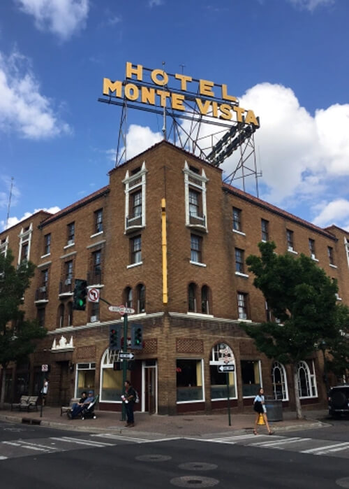 Four-story brick building in middle of picture with yellow Hotel Monte Vista sign at top. Blue sky with white clouds in background,