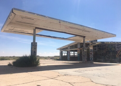 Abandoned gas station with large white canopy and empty repair shop and blue sky in background and cracked pavement below.