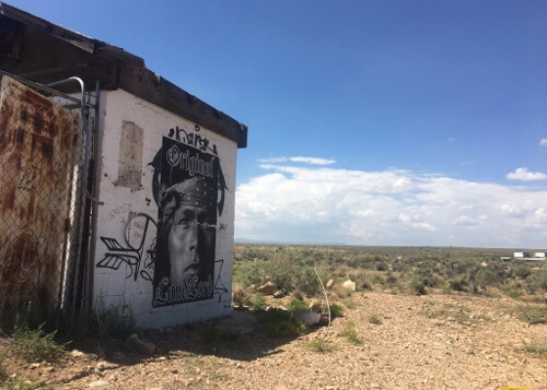 Face of a Native American painted on white wall of abandoned building at left, with brush covered desert disappearing into the white clouded blue sky horizon in the distance at right.