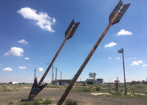 Two twenty-foot tall arrow looking structure sticking into the ground at an angle, with abandoned cement lot and blue sky with a few clouds in background.