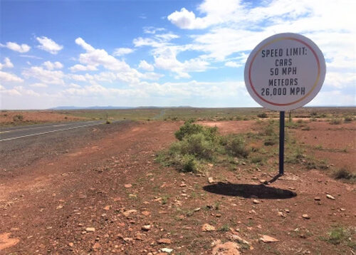 Round white sign reading "Speed Limit: Cars 50 mph, Meteors 26,000 mph" at right, with red desert sand and some green brush on the ground. Blue sky with white clouds in background.