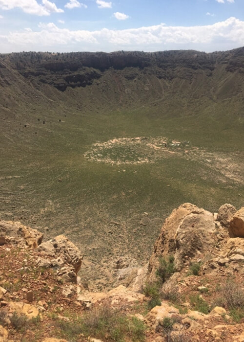 Massive "bowl" shaped crater in the desert terrain filled with green desert brush and some yellow rock at bottom foreground. Blue sky with clouds in background.