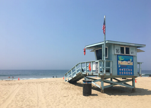 Aqua-colored lifeguard tower on yellow-sand beach with American flag, calm ocean and blue sky in background, light beige sand below.