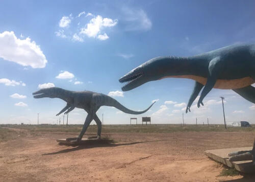 Two identical dinosaur statues with slender bodies and long extended necks, sideview, on desert dirt below and blue sky and scattered clouds above.