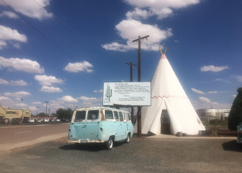 Late 1960's style Volkswagen blue van parked in front of white teepee in middle, with pavement at bottom and blue sky with scattered clouds above.