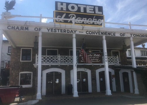 Two-story adobe brick hotel with white wooden support beams and white framed door entries, sign at top reading "Hotel El Rancho" in white lettering and blue sky above. 