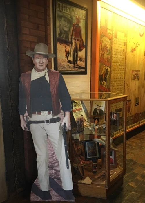 Cardboard life-size figure of John Wayne wearing cowboy outfit and cowboy hat at left, and a display case of movie memorabilia at right against a wall of movie posters.