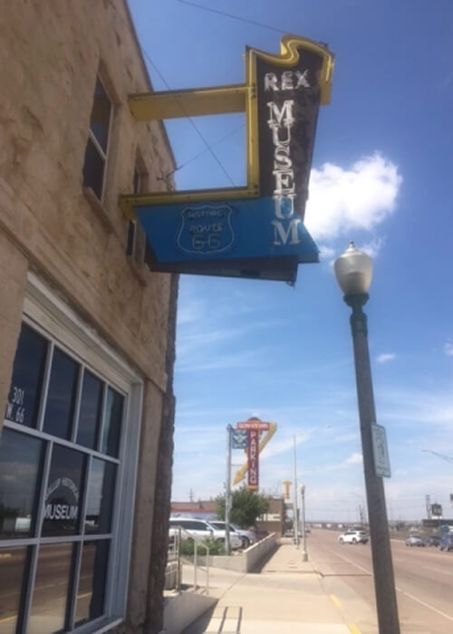 Adobe brick building at left, with window on first floor and adobe brick on second floor with neon sign sticking out overhead saying "Rex Museum." At right is street disappearing into horizon and street lamp, with blue sky and scattered clouds above.