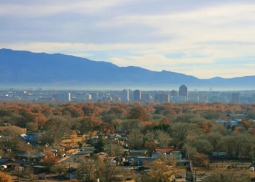 Series of downtown five to twenty-story buildings in the distance in middle of photo against a backdrop of dark mountain ridges, with green and orange trees below and hazy blue sky with clouds above.