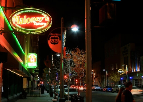 Night photo with neon in yellow lettering and green border reading "Maloney's" at upper left. At right is the street lined with cars and white and blue street lights.