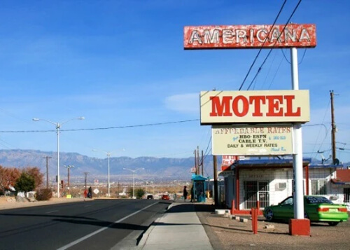 Rusty red sign at upper right reading "Americana" and newer looking sign below in red lettering reading "Motel. At left is street disappearing into mountain ridge horizon, blue sky above.