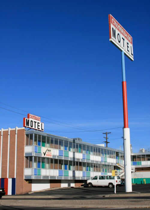 White and red sign reading "Motel" at right with white, red and blue post. Two story motel at left with alternating boxes of white, green and blue lining the walkways and rooms. 