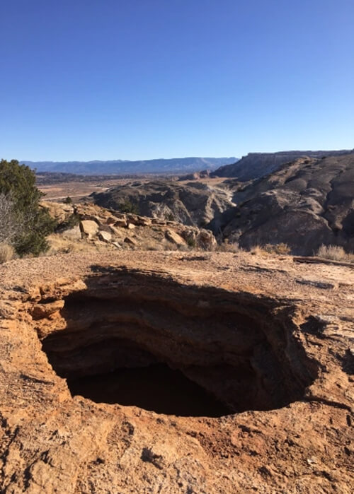 Circular black hole in a section of hard red rock, with some mountain ridges in background and blue sky above.