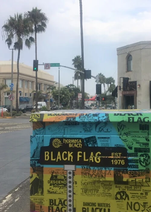 Sticker-covered postal box at bottom displaying various rock bands, with the largest sticker reading "Black Flag". Two-story light brown buildings, palm trees and grey sky in background.