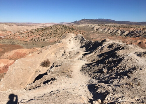 White, grey and reddish dirt and rock terrain along some mountain ridge tops with a faint trail at middle for hiking or biking. Blue, cloudless sky above.