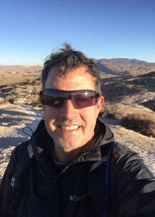 Close up picture of author Mark Loftin wearing dark jacket and sunglasses, hair being blown in wind, with some white sand mountains in background. Blue sky above.