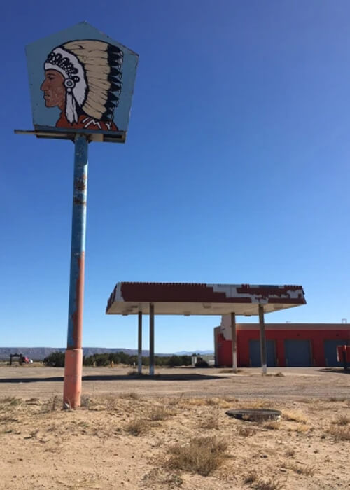 Abandoned gas station with sign at upper left of Native American wearing feathers and canopy with decayed red paint in middle. Garage building in far background and blue sky above.