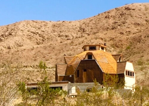 Half-dome shaped home with tan roof against backdrop of light brown dirt and rock mountain. Home has barb wire fence around it.