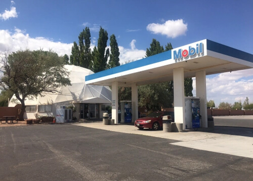 Mobil gas station at right with half-dome shaped food market behind it at left, black pavement at bottom and blue sky with a few white puffy clouds behind it.