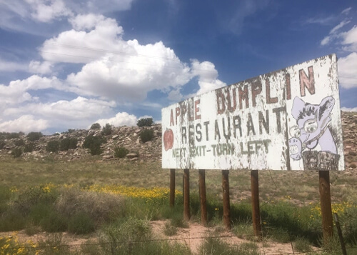 Wooden white painted billboard with faded and cracked paint saying "Apple Dumplin Restaurant", with some green desert brush and yellow flowers below, and blue sky with puffy white clouds above.