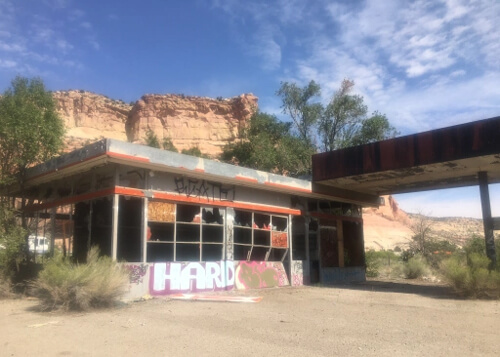 Abandoned gas station with empty window frames and cracked paint and graffiti, with red rock ridge behind and blue sky with some scattered clouds above.