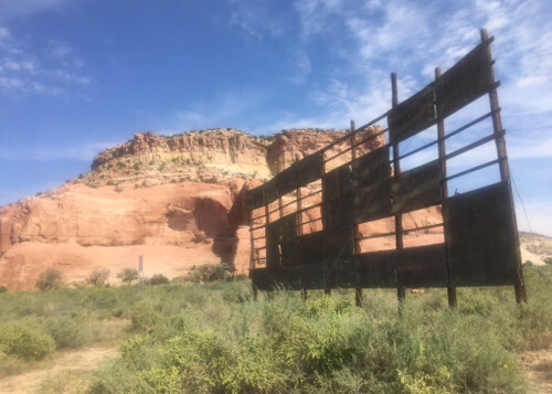 Wooden frame fragments of old billboard, with green desert brush at bottom and a red rock hill behind under a blue sky with a few scattered clouds.