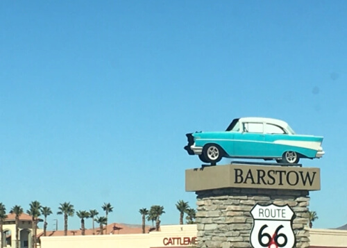 Sign of Barstow at right, with model 1950's car on top of sign and Route 66 sign below the sign. Palm tree tops and blue sky in background.