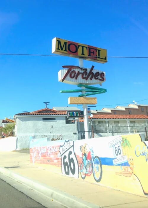 Yellow and orange box-design motel sign, with cursive sign underneath reading "Torches" and green arrow underneath point right. Two story motel with light blue paint and red roof in middle and right, with Route 66 painted brick wall below. Bright blue sky in background.