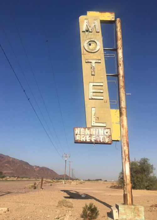 Rusty yellow neon sign displaying "Motel" standing tall in phone by itself on dirt lot with blue sky in background.