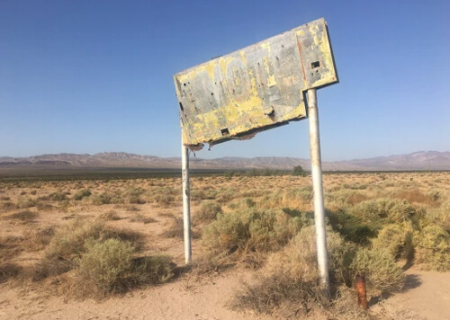 Rusty yellow sign with illegible lettering standing by itself on sand covered with desert bushes, with mountain ridge and blue sky in background.