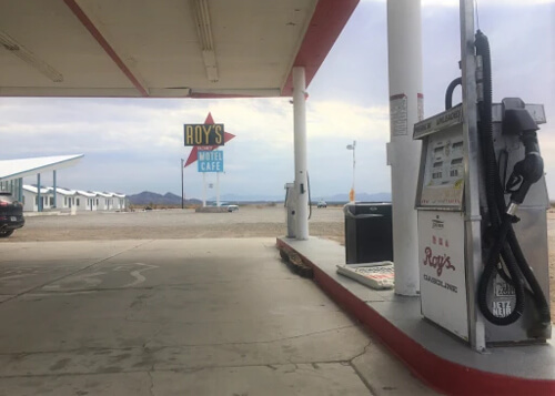 1970's style gas pump at bottom right underneath flat gas station canopy. Roy's sign in the distance against a grey cloudy backdrop.