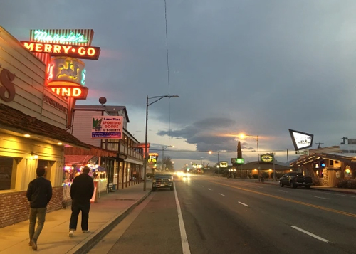 Row of single story businesses and shops on both sides of street disappearing straight into the horizon, under a cloudy sky at sundown.  Bright red neon sign at upper left, saying "Merry Go."