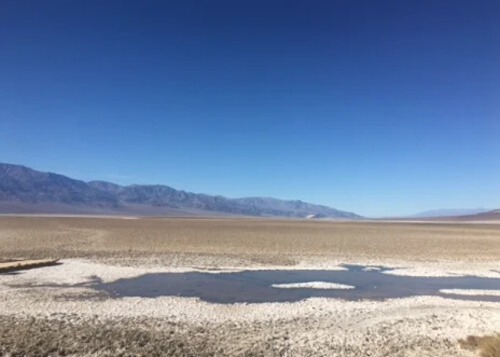 Dark brown mud pond in foreground, surrounded with some white salt. Thin range of dark brown mountains in middle. Blue sky with no clouds in background. 