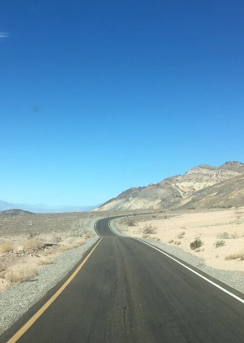 Thin, paved, single-lane road goes straight then turns right and disappears into thin brush covered desert sand hills. Blue sky with no clouds in background. 