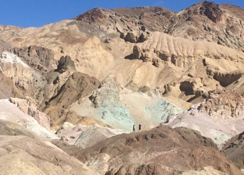 tacked jagged desert ridges of dark brown, light beige, bright white and green colored oxidized sand, with background of blue sky in top portion.