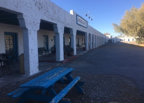 White-colored single-story building with arches extends in left side into the horizon, with blue bench in foreground and light brown leaved tree at far right. Blue sky fills in upper right portion.