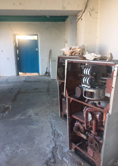 Remnants of two old gas pumps side by side at right in a white colored  corridor. In distance in middle right is a blue painted door, with the sun beaming through an opening at the top.