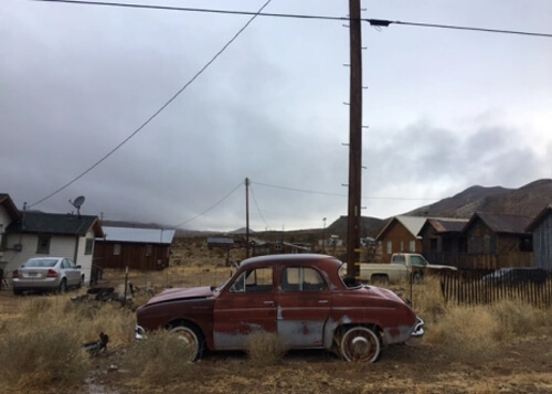 Old rusty 1930's red car with faded paint in foreground among some old wooden houses and yellow desert brush in foreground. Cloudy sky in background.