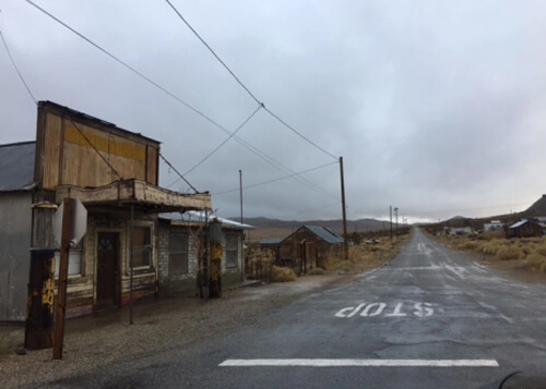 Uneven wet paved road disappearing into a grey, cloudy horizon on right side, and a couple of abandoned old wooden storefronts on left side.