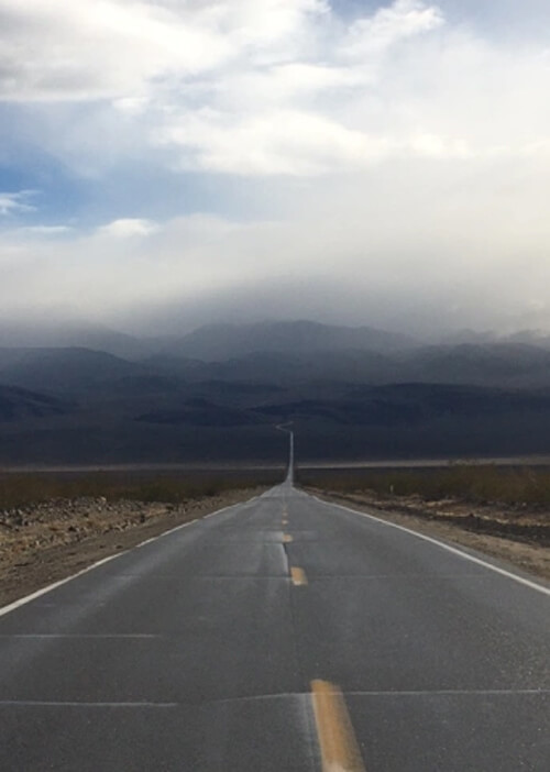 Old two lane highway with yellow stripes in middle gradually disappearing straight into desert mountains, blanketed with dark rain clouds.