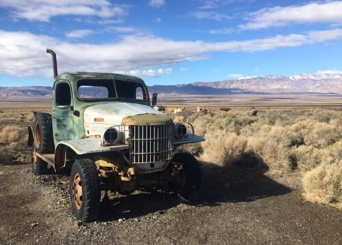 Abandoned old rusty truck with faded white hood and faded green cab on left side foreground. Yellow desert brush on right side. Blue sky with white cloud streaks in background.