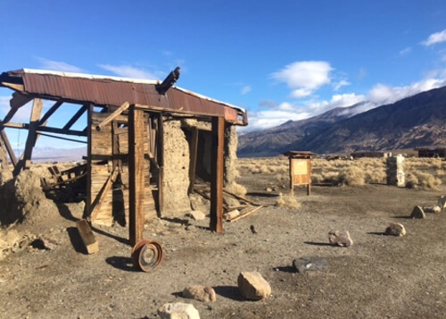 Remnants of old adobe brick building on left side foreground, with desert hills on right side and blue sky with puffy white clouds in background.