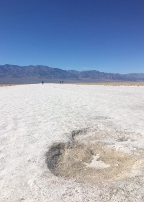 White salt desert ground with a few people far off in the distance in middle of photo. Thin range of dark brown mountains in middle, and blue sky with no clouds in background.