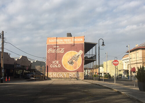 Mural of Coke sign and bottle on side of building in center of downtown area, some building on the right and left of it, grey cloudy sky above.