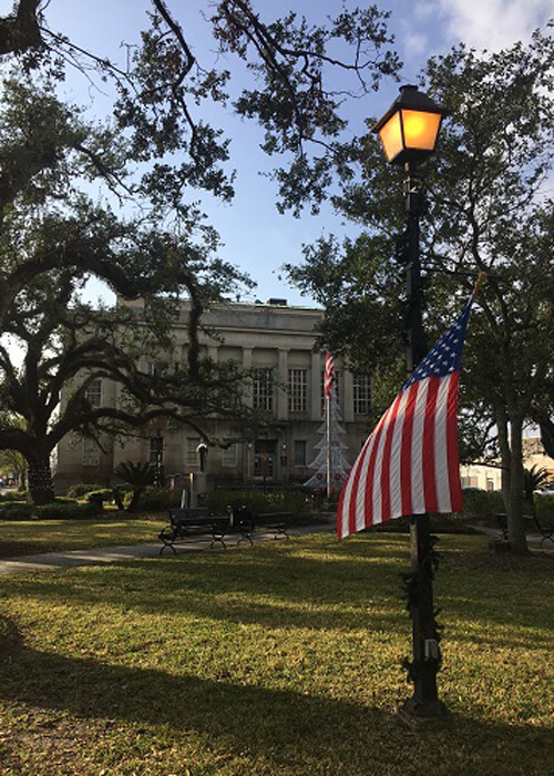 American flag hanging from lit lamp post, courthouse building in background. Blue sky with some clouds above.